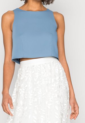 Kobieta ubrana w bezrękawnikowy niebieski top i białą spódnicę z haftem liści, widziana od ramion do talii.