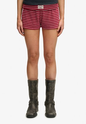 Persoon die rood-wit gestreepte shorts draagt met het Superdry-logo en donkergrijze kniehoge laarzen tegen een witte achtergrond.