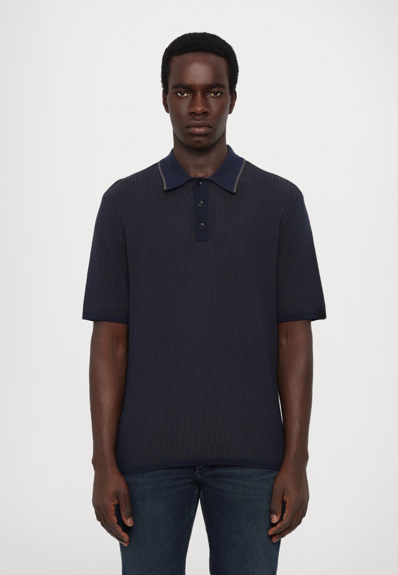 Mørk marineblå pol shirt med korte ærmer, ribbet texture, tre-knaps placket og kontrastfarvet krave. Bæres med mørke jeans.