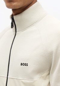 Κοντινό πλάνο ενός ατόμου που φοράει μπεζ πουλόβερ με φερμουάρ, με ριμπ γιακά και το λογότυπο "BOSS" στο στήθος.