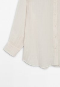 Chemise beige à manches longues avec texture subtile et poignet large à un seul bouton sur fond blanc uni.