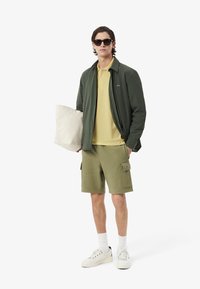 Groene lichte jas, gele polo, olijfgroene cargoshorts, witte sneakers en zwarte zonnebril. Houdt een crèmekleurige stoffen tas vast.