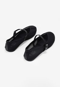 Bailarinas negras hechas de tela suave con punta redonda y una correa en el tobillo. Presentan una suela de goma texturizada y un logo sutil en la parte posterior.