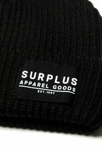 Crni pleteni zimski šešir s rebrastom teksturom. Ima pravokutnu crnu oznaku s natpisom "SURPLUS APPAREL GOODS" i "EST. 1997" u bijelom tekstu.