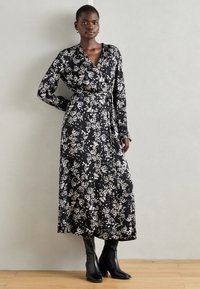 Crna cvjetna haljina s dugim rukavima, strukirana, s dužinom do gležnjeva. Tkanina ima bijele uzorke lišća. Crne čizme s visokom petom.