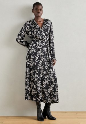 Svart blomstrete omslagskjole med lange ermer, innsvingt midje og ankel-lengtet skjørt. Stoffet har hvite bladmønstre. Svart hæl støvler.