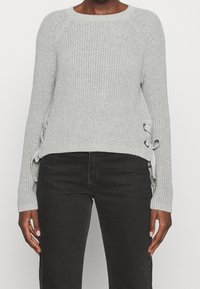 Jasnoszary sweter z dzianiny z okrągłym dekoltem, prążkowaną fakturą i ozdobnymi sznurowaniami po bokach. Długie rękawy, lekko skrócony fason.