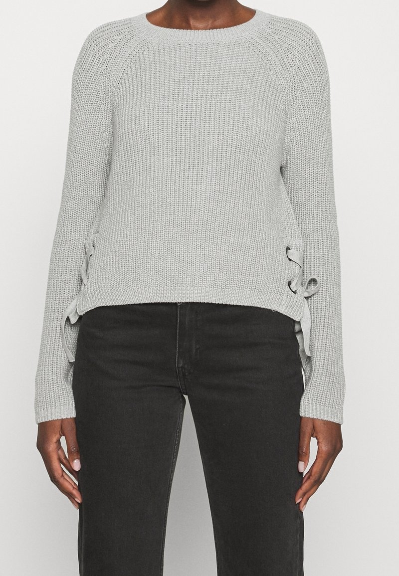Jasnoszary sweter z dzianiny z okrągłym dekoltem, prążkowaną fakturą i ozdobnymi sznurowaniami po bokach. Długie rękawy, lekko skrócony fason.