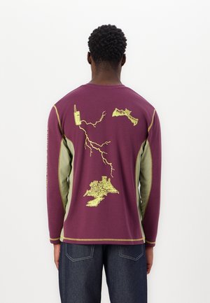 Homme portant un t-shirt à manches longues violet avec des panneaux latéraux verts et un motif jaune représentant des montagnes, des éclairs et un talkie-walkie dans le dos.