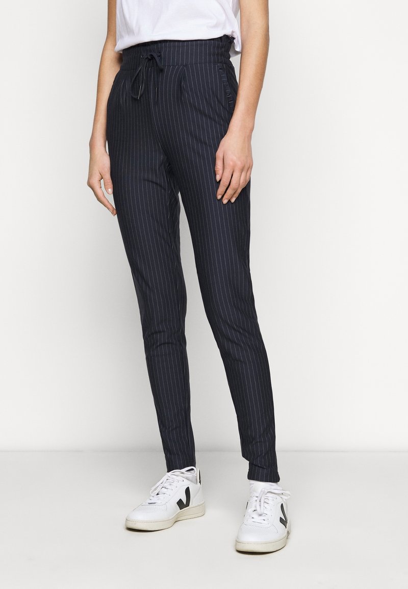 Personne portant un pantalon slim noir à fines rayures avec taille à cordon et des baskets blanches avec logo noir, debout devant un fond blanc.