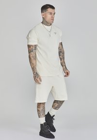 T-shirt corto a maniche corte di un beige chiaro e pantaloni corti abbinati, realizzati in tessuto morbido. Stivali neri e calze bianche. Tatuaggi visibili sulle braccia.