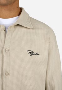 Beige waffle-textúrájú ing gallérral és gombos elülső résszel. A bal mellrészen fekete hímzett "Pegador" logó található.