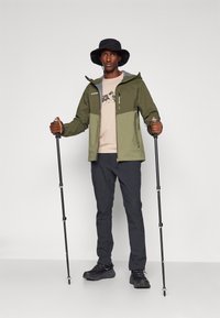 Olivengrøn vandtæt jakke med grå detaljer, beige T-shirt med bjergmotiv, sorte bukser og vandrestave i en minimal baggrund.