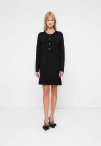 Μαύρο φόρεμα με μακριά μανίκια και στρογγυλή λαιμόκοψη, πέντε κουμπιά μπροστά και φαρδύ τελείωμα. Συνδυασμένο με μαύρες γόβες με τακούνι που έχουν λεπτομέρειες σε σχήμα φιόγκου.