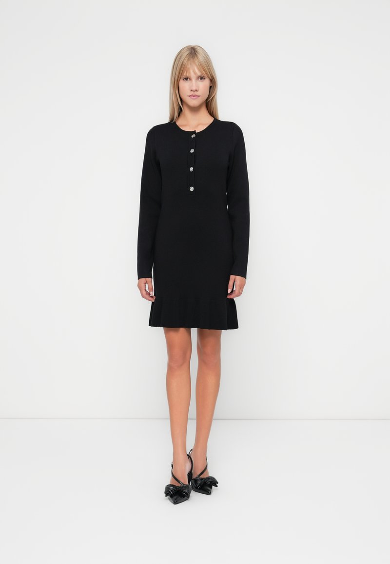 Μαύρο φόρεμα με μακριά μανίκια και στρογγυλή λαιμόκοψη, πέντε κουμπιά μπροστά και φαρδύ τελείωμα. Συνδυασμένο με μαύρες γόβες με τακούνι που έχουν λεπτομέρειες σε σχήμα φιόγκου.