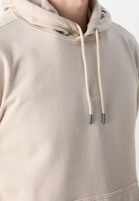 Beige hoodie i mjukt tyg, med dragsko med silvriga ändar och en subtil präglad logotyp på bröstet.