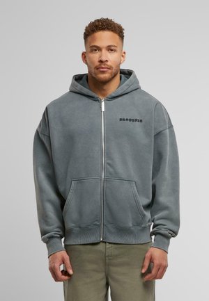 Gray zip-up hoodie with a front pocket, drawstring hood, and black logo text. Soft fabric with ribbed cuffs and hem. Styled with light pants.