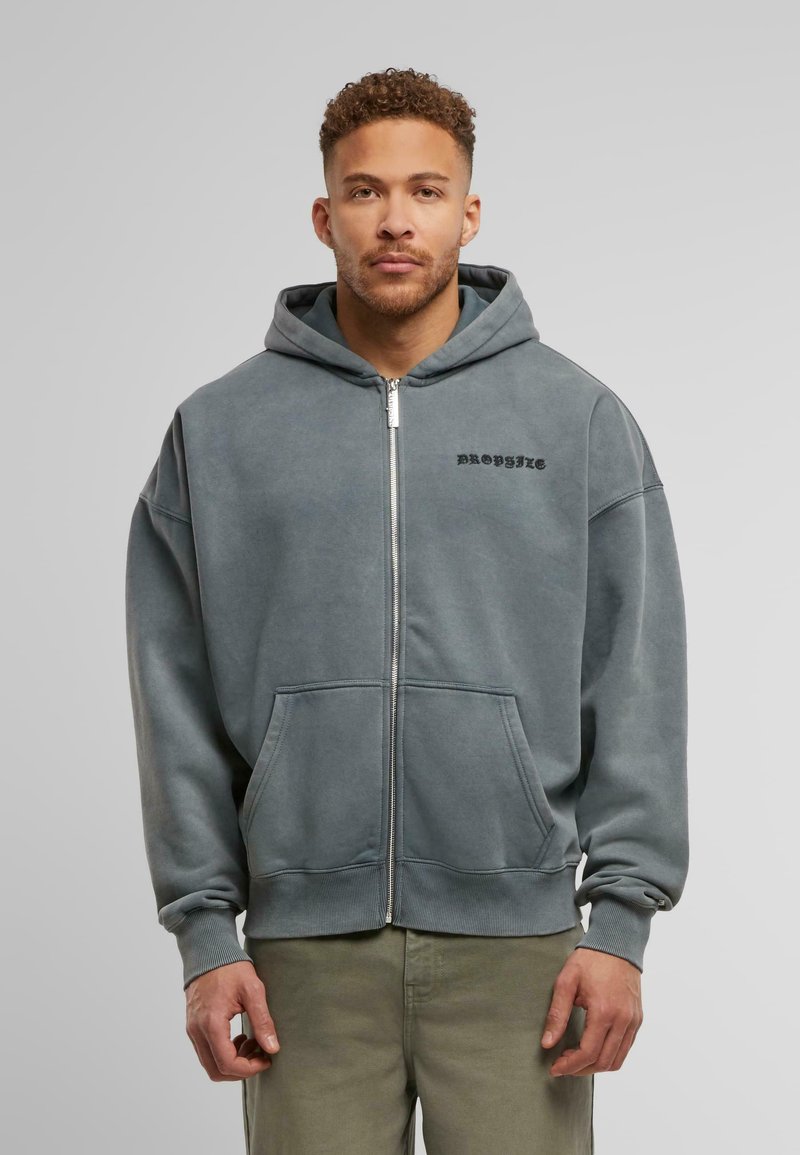 Grauer Reißverschluss-Hoodie mit einer Vorderseite-Tasche, Kapuze mit Kordelzug und schwarzem Logodruck. Weicher Stoff mit gerippten Bündchen und Saum. Kombiniert mit hellen Hosen.