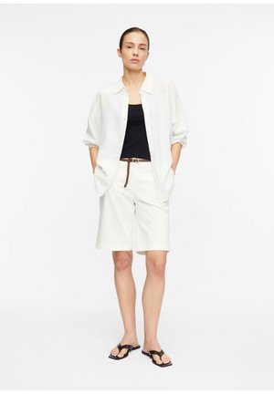 Kobieta stojąca, ubrana w białą koszulę, czarny top na ramiączkach, białe krótkie spodenki do kolan z brązowym paskiem i czarne sandały na białym tle.