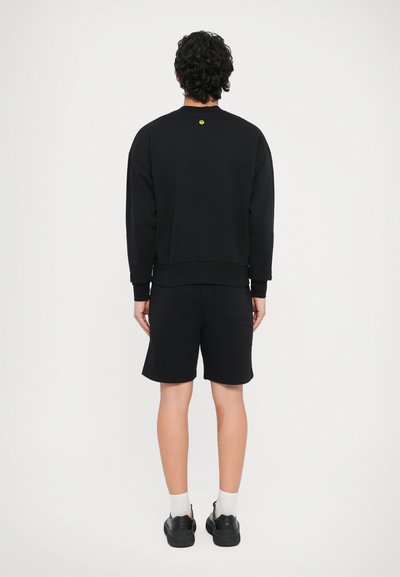 Melna sporta jaciņa ar apaļu apkakli un garām piedurknēm, kombinēta ar melnām šortām. Atpakaļpusē neliels dzeltens logotips. Gluda tekstūra.