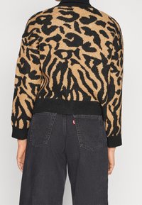 Person trägt einen braun-schwarzen Tiermotiv-Pullover mit schwarzen Rippbündchen und schwarze hochgeschnittene Jeans mit sichtbaren Gesäßtaschen.