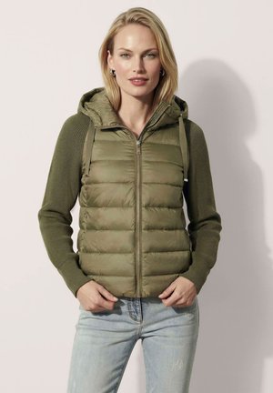 Blonde Frau trägt eine olivgrüne gesteppte Weste mit Kapuze und gestrickten Ärmeln, kombiniert mit hellblauen Jeans, steht vor einfachem Hintergrund.