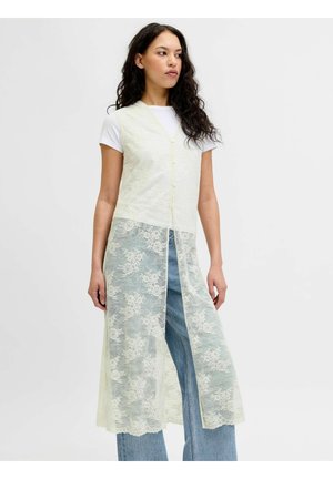 Vrouw draagt wit kortärmelig topje, blauwe spijkerbroek en een lange, mouwloze crèmekleurige kanten vest met bloemenpatroon, staand tegen een effen achtergrond.