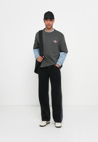 Uomo con berretto nero, felpa grigia con logo rosa, maniche di camicia azzurra chiara, pantaloni neri, sneakers bianche e borsa a tracolla nera su sfondo bianco.