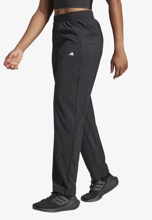 Persoon die zwarte Adidas trainingsbroek en zwarte Adidas sneakers draagt, getoond van taille tot voeten tegen een witte achtergrond.
