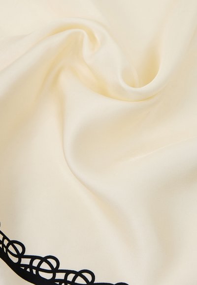 Tissu de couleur crème avec une texture lisse et des plis subtils, présentant un motif ornemental noir partiel dans le coin inférieur gauche.