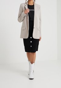 Ljusbeige skräddarsydd kavaj med två framfickor, bärs över en svart skjorta med djärv vit text, tillsammans med en svart pennkjol och vita stövlar.