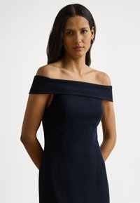 Μπλε σκούρο φόρεμα off-the-shoulder με εφαρμοστή σιλουέτα, απαλό ύφασμα και διακριτικές ραφές, αναδεικνύοντας έναν κομψό και μίνιμαλ σχεδιασμό.