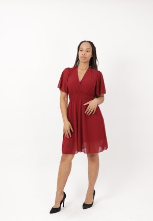 Donna con i capelli intrecciati che indossa un vestito rosso al ginocchio e tacchi alti neri, in piedi davanti a uno sfondo bianco semplice.