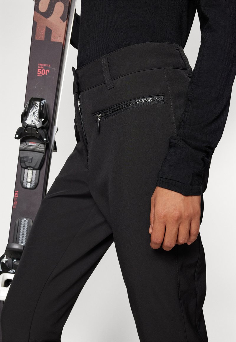 Schwarze Skihosen mit glatter Textur, ausgestattet mit einer Reißverschlusstasche und einer taillierten Passform. Ein Ski ist teilweise im Hintergrund zu sehen.