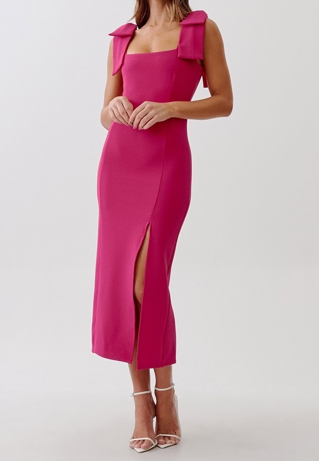 Femme portant une robe midi rose ajustée sans manches avec de grands nœuds aux épaules et une fente haute sur la cuisse, associée à des talons blancs à lanières.