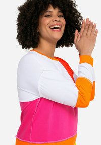 Barevná mikina s bílými, oranžovými, růžovými a červenými částmi. Tkanina má hladkou strukturu a kontrastní švy.