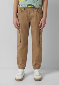 Beige cargobroek met een elastische tailleband, zijzakken en taps toelopende boorden, gecombineerd met witte sneakers met lichtbruine zolen.