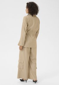Beiger maßgeschneiderter Blazer mit taillierter Passform, kombiniert mit weit geschnittenen Cargo-Hosen mit seitlichen Taschen. Glatte Textur und minimalistisches Design.