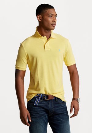 Kollane polo särk, millel on krae, lühikesed varrukad ja väike sinine logo. Ühendatud tumedate teksadega ja pruuni vööd, millel on triibulised aktsendid.