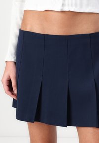 Navy pleated mini skirt made from woven fabric, featuring a fitted waistband and stitched detailing, shown with a white ribbed top.