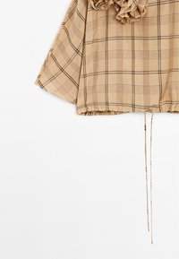 Beige geruit top met uitlopende mouwen, geplooide zoom en ruches. Heeft dunne zwarte en bruine lijnen in het patroon, en verstelbare trekkoorden.