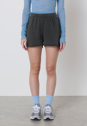 Person iført sorte shorts, blåstribet langærmet skjorte, lyseblå sokker og grå sneakers, stående på en betongulv mod en hvid væg.