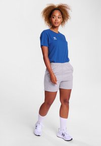 Blauwe katoenen T-shirt met ronde hals, gecombineerd met lichtgrijze elastische shorts. Het model draagt witte sokken en sneakers en staat tegen een effen achtergrond.