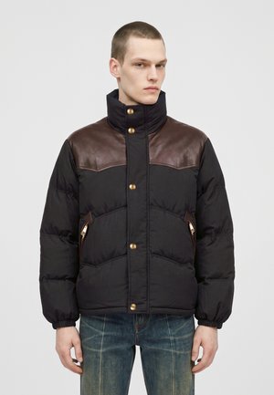 Un mannequin masculin porte une doudoune noire avec un empiècement en cuir marron sur la poitrine, un col montant, des boutons dorés et un jean bleu.