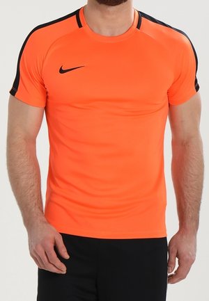 Mężczyzna noszący jaskrawy pomarańczowy sportowy t-shirt z krótkim rękawem z czarnymi paskami na rękawach i czarnym logo Nike na piersi, w połączeniu z czarnymi szortami.