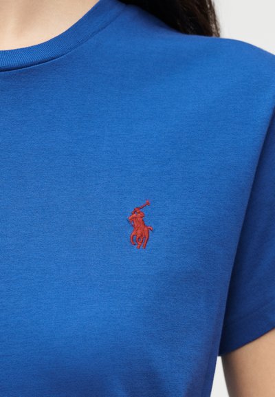 Κοντινό πλάνο ενός ατόμου που φοράει μπλε μπλούζα με στρογγυλή λαιμόκοψη και μικρό κόκκινο κεντητό λογότυπο παίκτη πόλο στο στήθος.