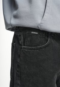 Schwarze Denim-Jeans mit einer verblassten Textur, ausgestattet mit einer Seitentasche und einem kleinen "PEGADOR"-Label in der Nähe des Bundes. Mit einem grauen Hoodie kombiniert.
