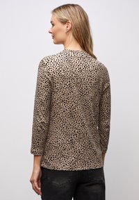 Beige-Oberteil mit schwarzem Punktmuster, langen Ärmeln, rundem Halsausschnitt und glatter Textur. Die Rückansicht zeigt eine lockere Passform und ein schlichtes Design.