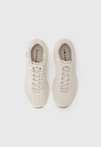Beige athletic shoes with a textured mesh upper, flat laces, and a supportive sole, featuring branding on the tongue and side accents.