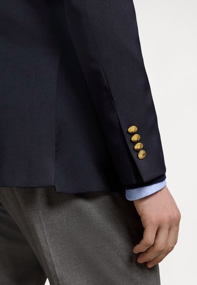 Blazer azul-marinho com botões dourados, corte ajustado, e calças cinzentas. Camisa azul-claro visível na manga, exibindo tecido texturado.